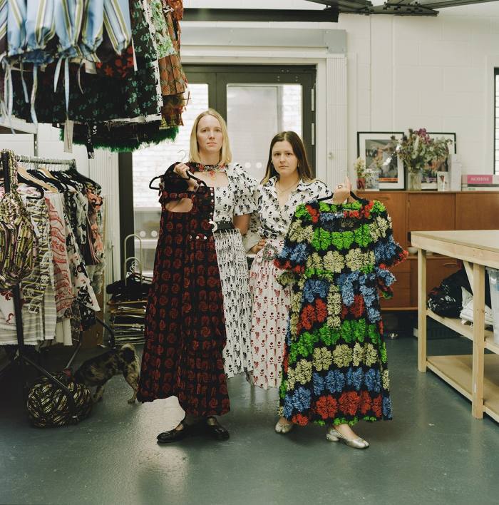 Laura Lowena and Emma Chopova, wearing printed midis beside a rack of clothes, hold up colourful ones in hangers