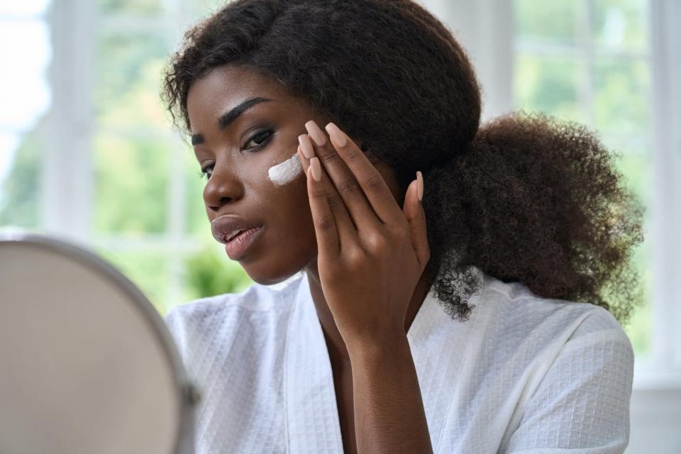 Attractive young black African woman model touching healthy flawless radiant smooth face skin care looking in bathroom mirror applying facial cream. Ethnic beauty skincare morning routine concept