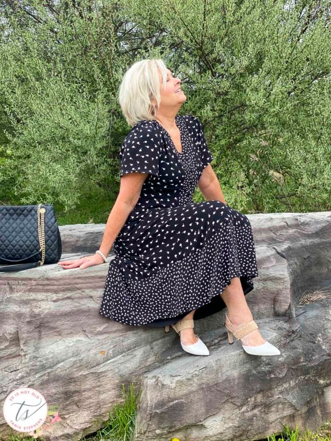 Fashion Blogger 50 Is Not Old is sitting on a rock wearing a black and white print dress from Talbots with beige and white heels
